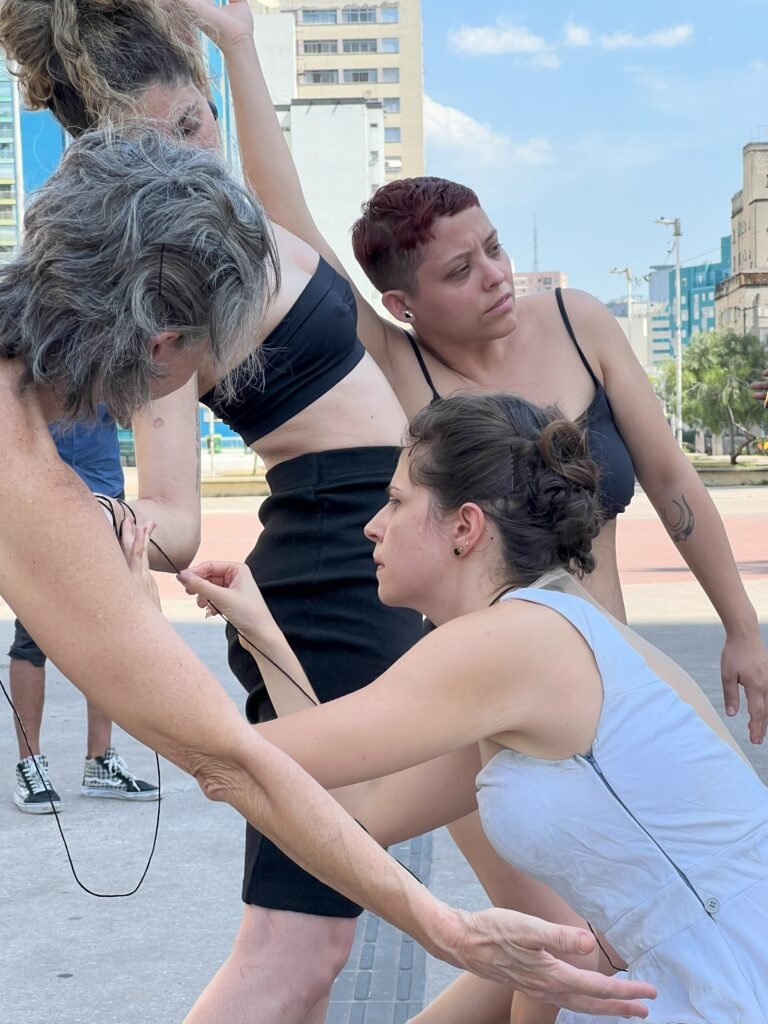 Performers se inclinam e se apoiam umas nas outras, ligadas pelo fio de sER.á.linha_do, na Praça Roosevelt