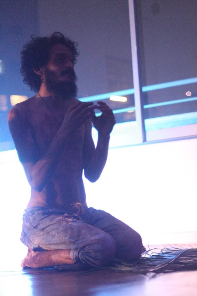 Ismael Trabuco ajoelhado, sem camisa, com as mãos juntas diante do peito, em luz azulada
