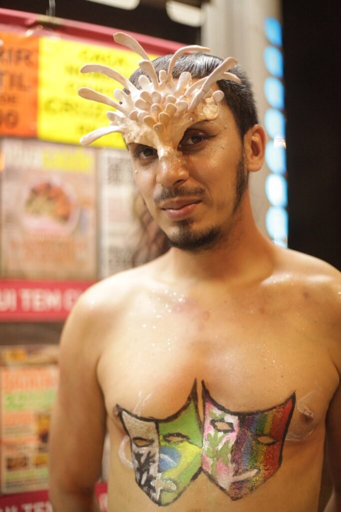 Performer com máscara esculpida sobre o rosto e máscaras de teatro pintadas com as cores do arco-íris no peito, no Largo do Arouche