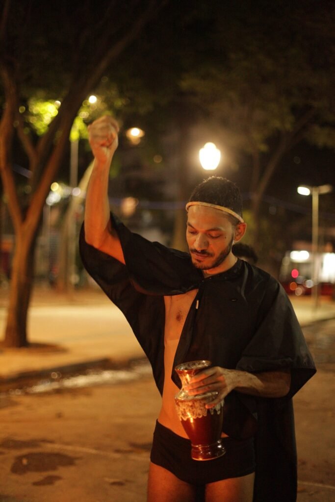 plexoisaac, de capa preta, segura o Vaso dos Desejos e ergue o outro braço no Largo do Arouche, à noite