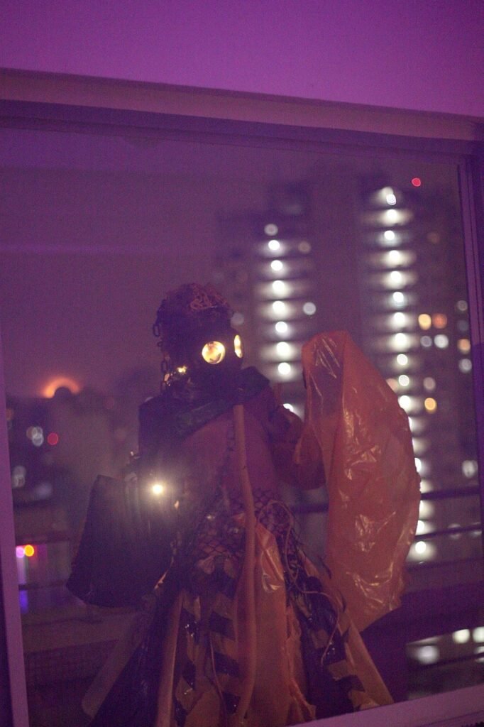Performer do Coletivo Metanoia com máscara de proteção e figurino de plástico, diante da janela com as luzes da cidade