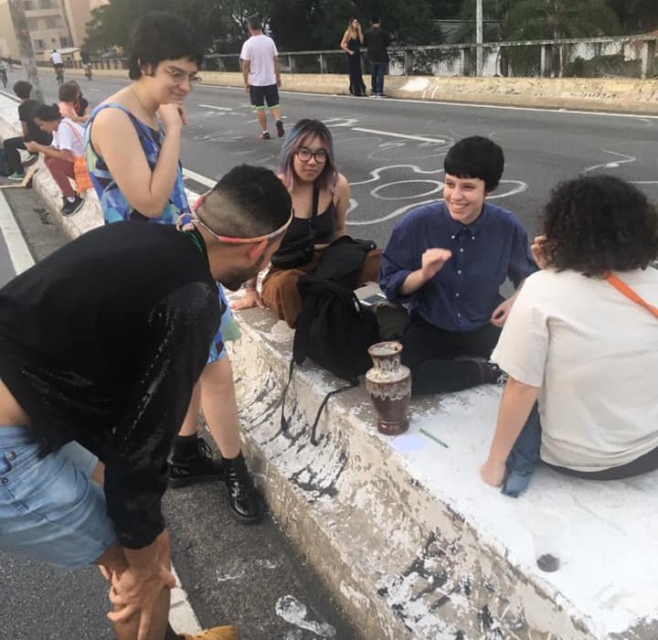 plexoisaac se inclina diante de pessoas sentadas na mureta do Minhocão; ao lado delas, o Vaso dos Desejos