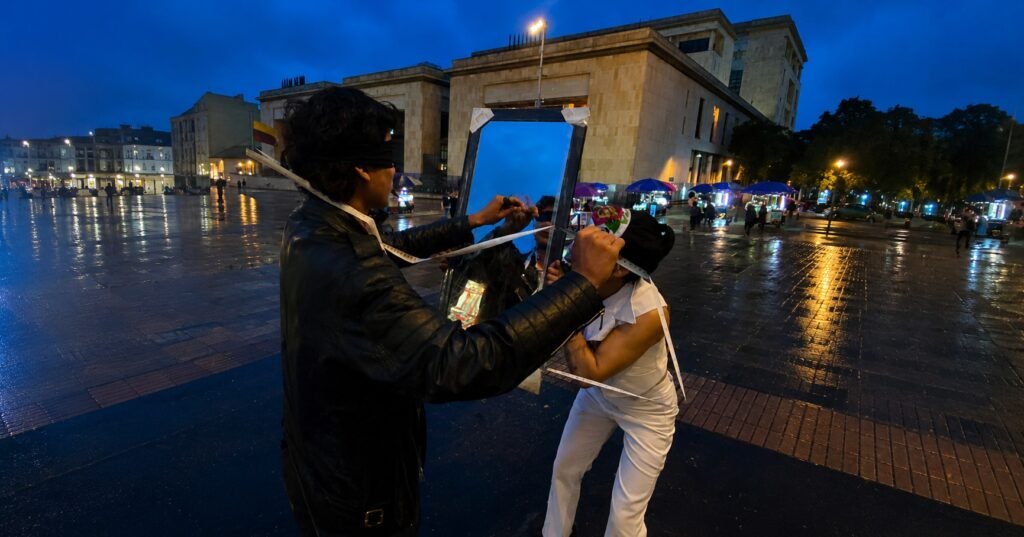 Dois performers numa praça molhada à noite, em Bogotá: um, de costas e de jaqueta de couro, segura uma fita métrica diante de um espelho; o outro, de branco, se curva ao lado