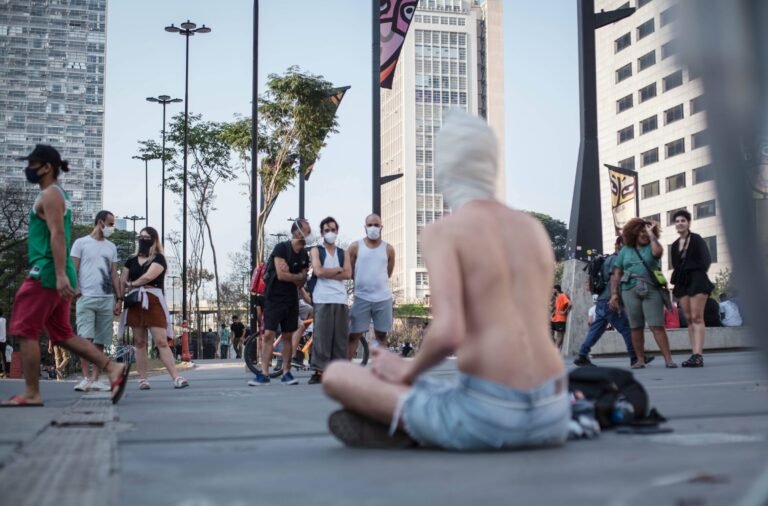 Reyan Perovano, de cabeça envolta em tecido branco, sentado no chão do Anhangabaú, observado pelo público