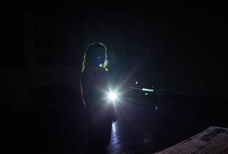 Integrante da Coletiva Transperformática Ruidosa Alma iluminada por trás por uma lanterna, em sala escura na Casa do Povo