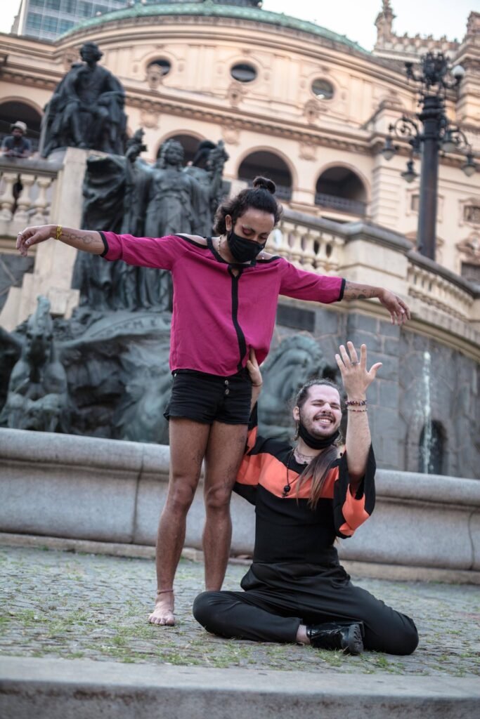 Performer da Ruidosa Alma de braços abertos diante da fonte do Theatro Municipal, com outra integrante sentada ao pé