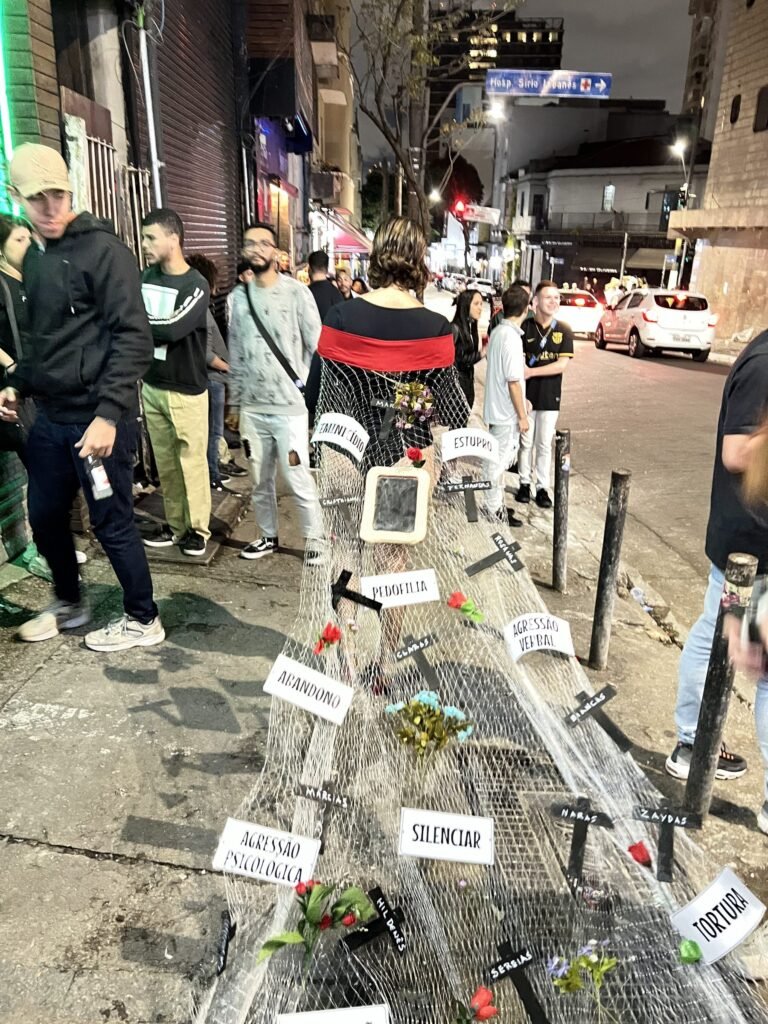 Cassia Pires, de costas, caminha pela calçada da rua Augusta arrastando uma rede presa ao corpo, com cruzes pretas, flores e placas com palavras como abandono e pedofilia; pessoas observam ao redor