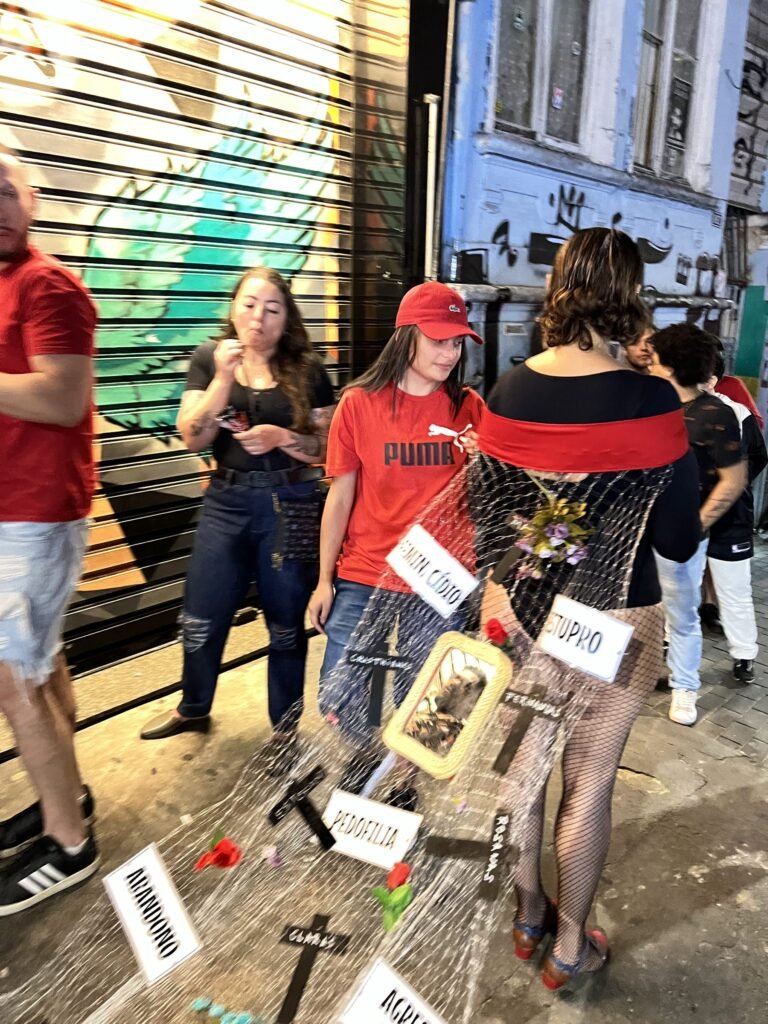 Cassia Pires, de costas, com a rede presa ao vestido e placas com as palavras feminicídio e estupro, passa entre pessoas na calçada