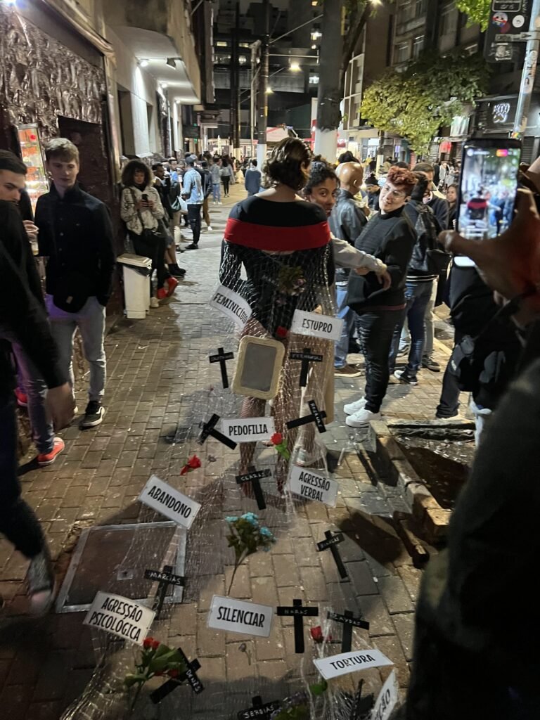 A rede de Arrasto se estende pela calçada atrás de Cassia Pires, com cruzes pretas, flores e placas com as palavras feminicídio, estupro, pedofilia, abandono e agressão verbal, em meio ao público da rua Augusta à noite