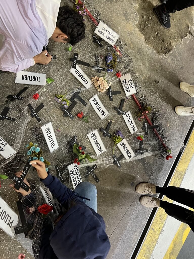 Vista de cima da rede estendida no chão, com cruzes pretas, flores e placas com palavras como tortura, covardia e silenciar