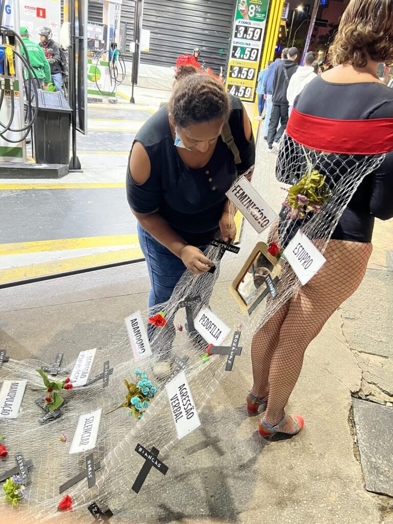 Uma mulher se agacha para mexer na rede presa ao vestido de Cassia Pires, diante de um posto de combustível