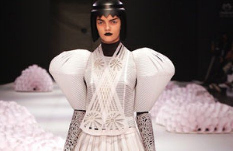 Modelo com cabelo preto em forma de capacete e vestido branco de papel recortado, com mangas volumosas, na passarela
