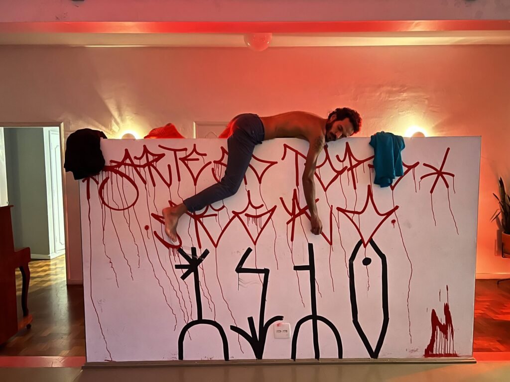 Ismael Trabuco deitado sobre uma divisória branca pintada com a palavra IsTo e respingos vermelhos, em sala iluminada de vermelho