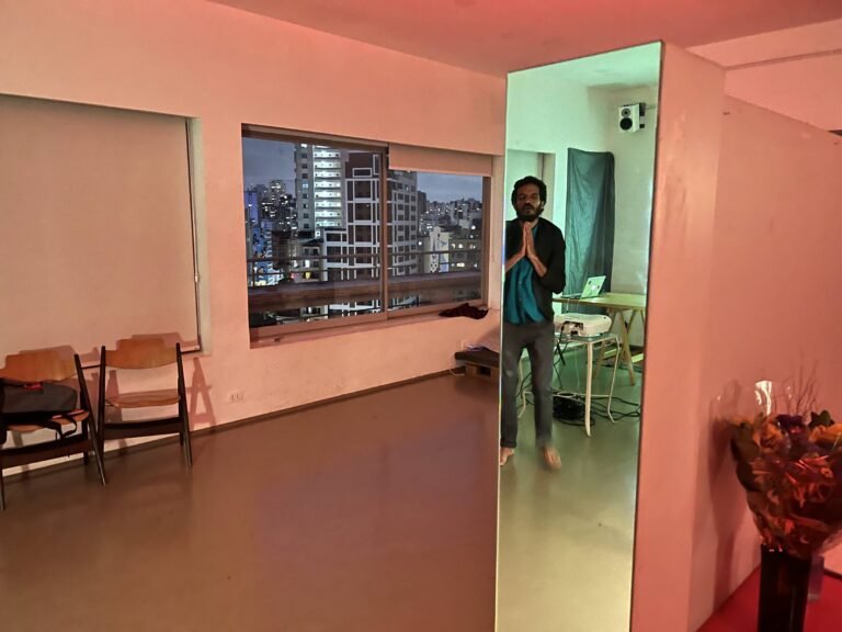 Ismael Trabuco, de mãos juntas, refletido num espelho da sala do Esponja, com a janela para os prédios do centro