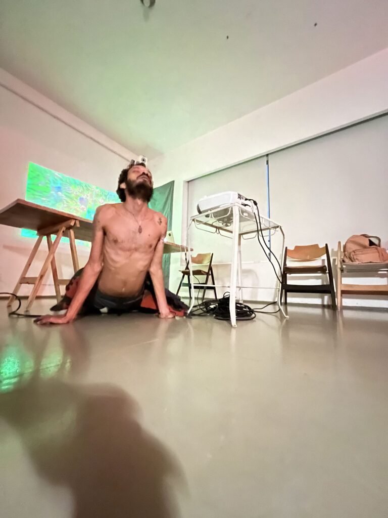 Ismael Trabuco, sem camisa, sentado no chão com o rosto voltado para cima, diante de uma projeção verde