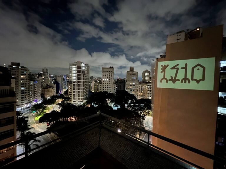 O nome IsTo projetado na empena de um prédio vizinho, visto do terraço do Esponja à noite, sobre o centro de São Paulo