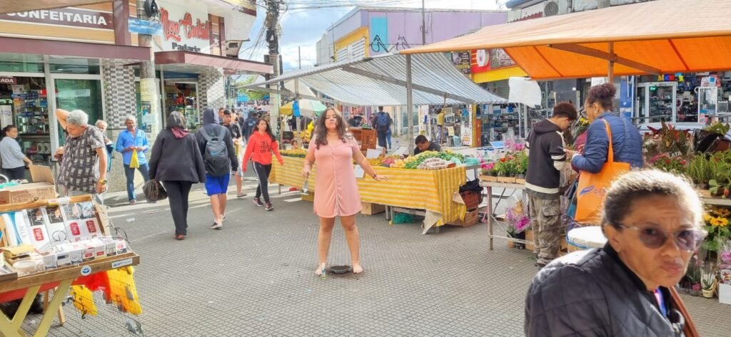 Wilka Sales, de vestido rosa, caminha entre as barracas de uma feira de rua no Itaim Paulista