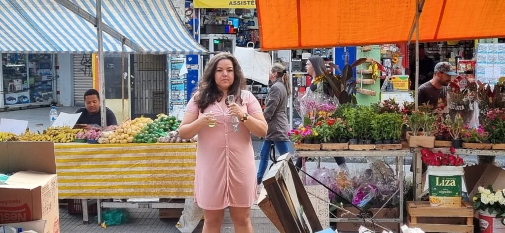 Wilka Sales, de vestido rosa, segura um copo diante das bancas de frutas e flores da feira