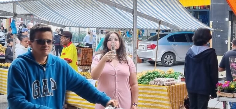 Wilka Sales bebe de um copo entre as barracas da feira, com pessoas passando em primeiro plano