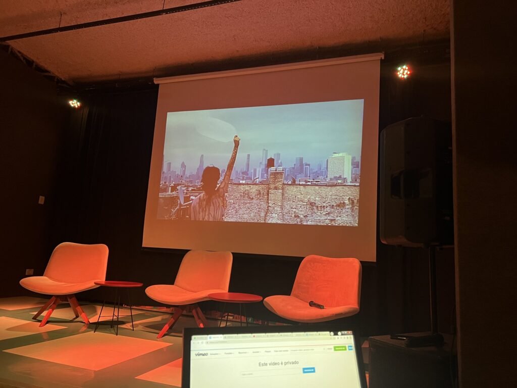 Auditório da Ação Educativa na noite de abertura: na tela, Hot Air, do coletivo Unides contra a colonização — uma pessoa ergue um balão transparente diante de prédios