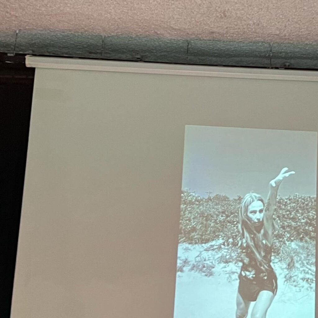 Videoperformance em preto e branco projetada na noite de abertura: mulher de cabelos longos ergue o braço entre a vegetação