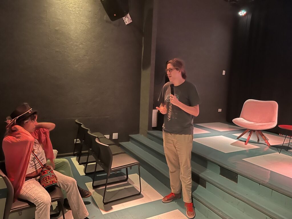 Artista fala ao microfone na conversa da noite de abertura, no auditório da Ação Educativa; ao lado, uma pessoa de chapéu e xale vermelho escuta