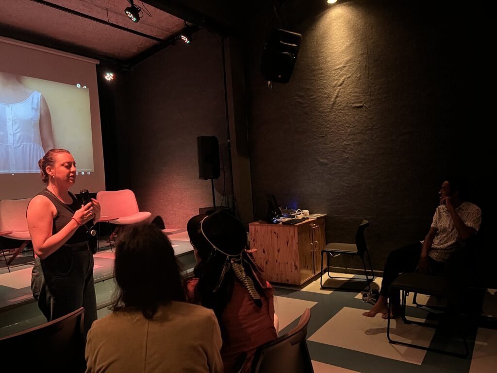 Artista fala ao microfone diante da tela, na conversa da noite de abertura, com o público sentado no auditório da Ação Educativa
