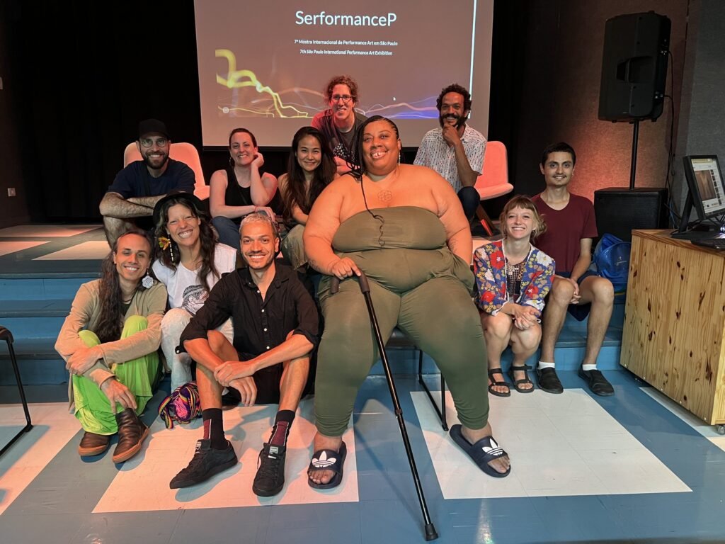 Artistas e equipe posam juntos no palco do auditório da Ação Educativa, diante da tela com a marca da SerformanceP, no fim da noite de abertura