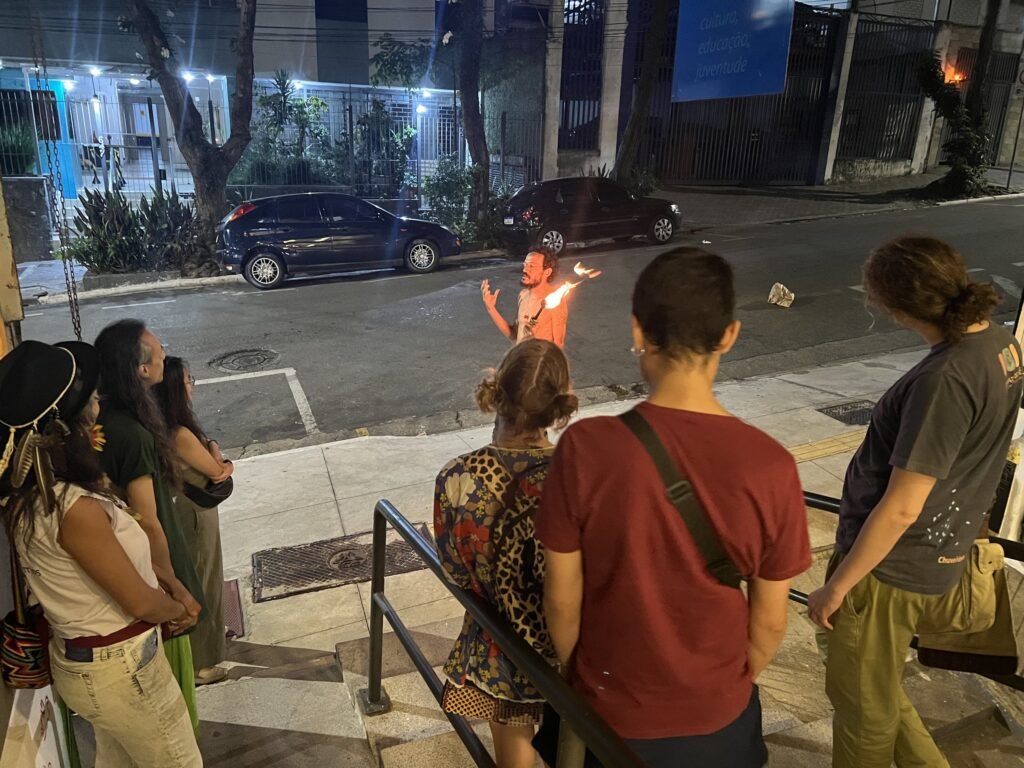 Público reunido na escada de entrada da Ação Educativa assiste a Ismael Trabuco com uma tocha acesa na rua, à noite