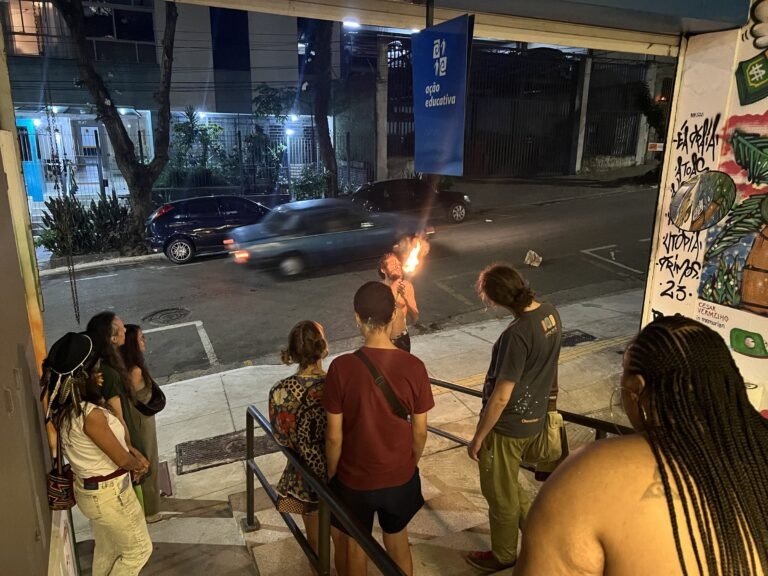 Ismael Trabuco com uma tocha acesa na rua enquanto um carro passa; o público observa da entrada da Ação Educativa