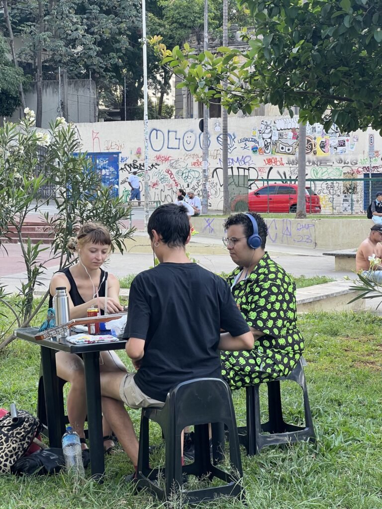 Jana Nunčič e Luan Gonçalves à mesa de Cycle of things, no gramado da Praça Roosevelt, com uma pessoa do público de fones de ouvido; ao fundo, muro grafitado