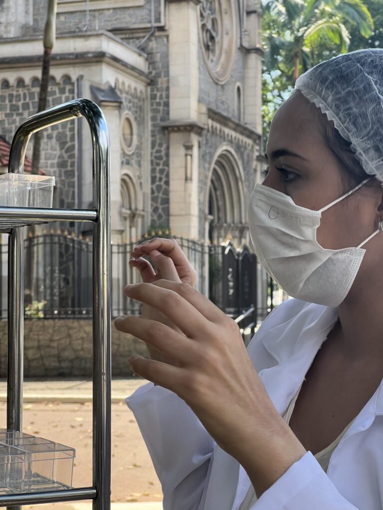 Marie Araújo, de máscara e touca, observa uma caixa transparente no carrinho de aço, diante da igreja