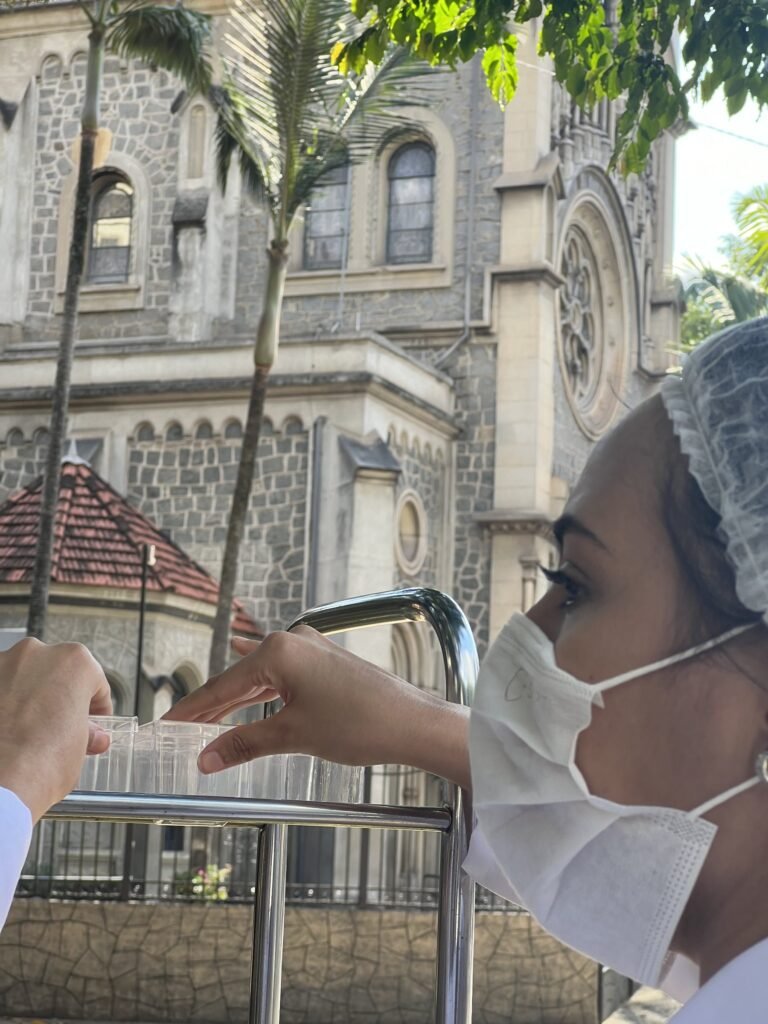 De perfil, Marie Araújo, de máscara e touca, segura uma caixa transparente junto ao carrinho de aço