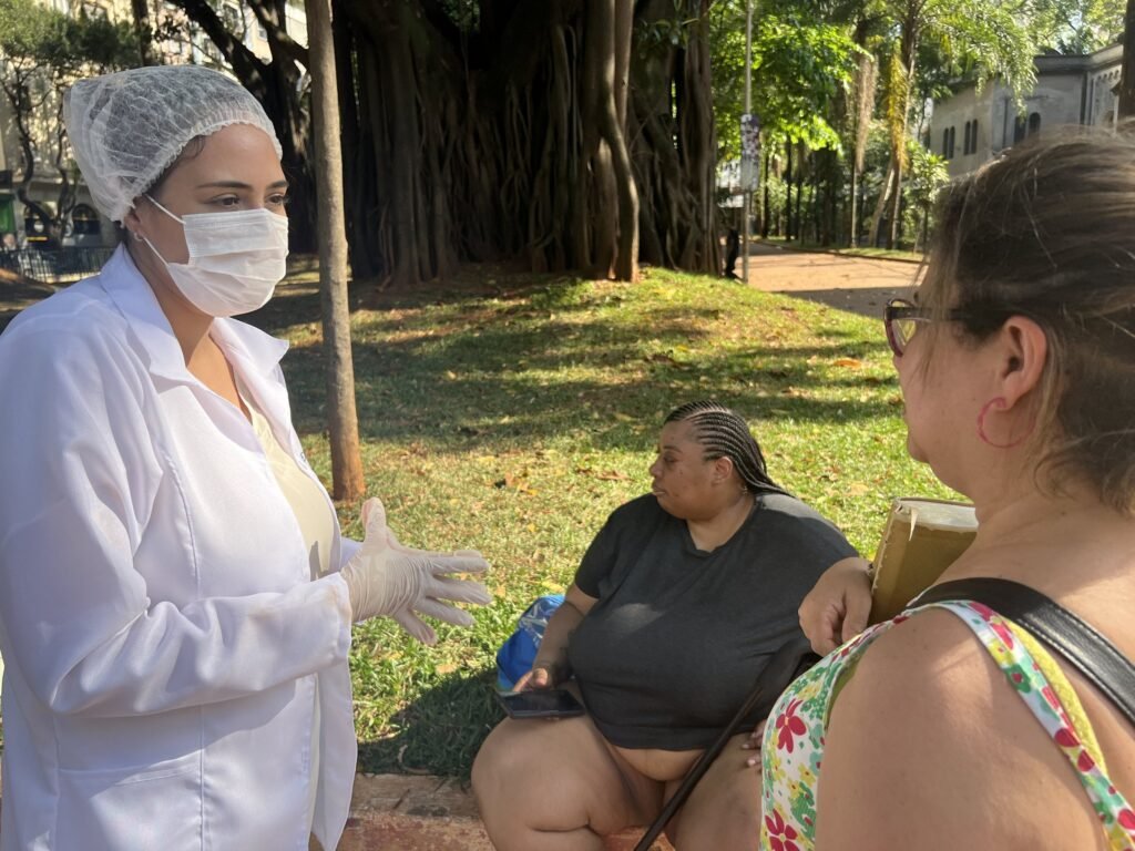 Marie Araújo, de jaleco e luvas, conversa com duas mulheres sentadas no gramado da Praça Roosevelt