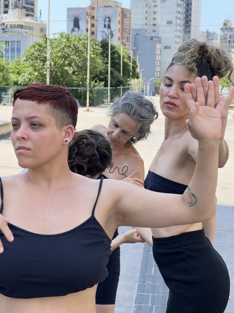 Performers de sER.á.linha_do de pé na Praça Roosevelt, com palavras escritas em fio preto sobre a pele; prédios ao fundo