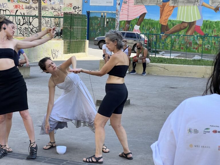 Uma performer de vestido claro se inclina e outra estende a mão, ligadas pelo fio preto, diante dos murais da Praça Roosevelt