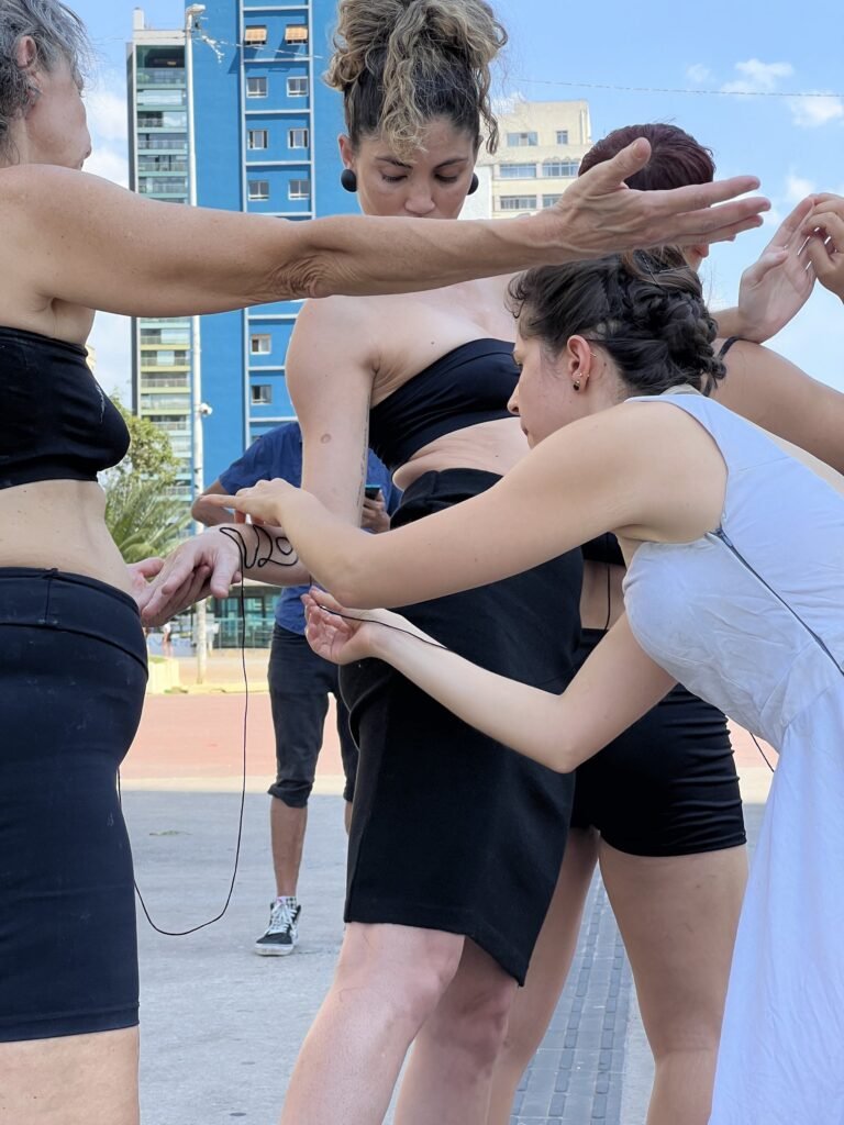 Uma performer de vestido claro estende o fio entre os corpos das colegas, sob o céu azul da Praça Roosevelt