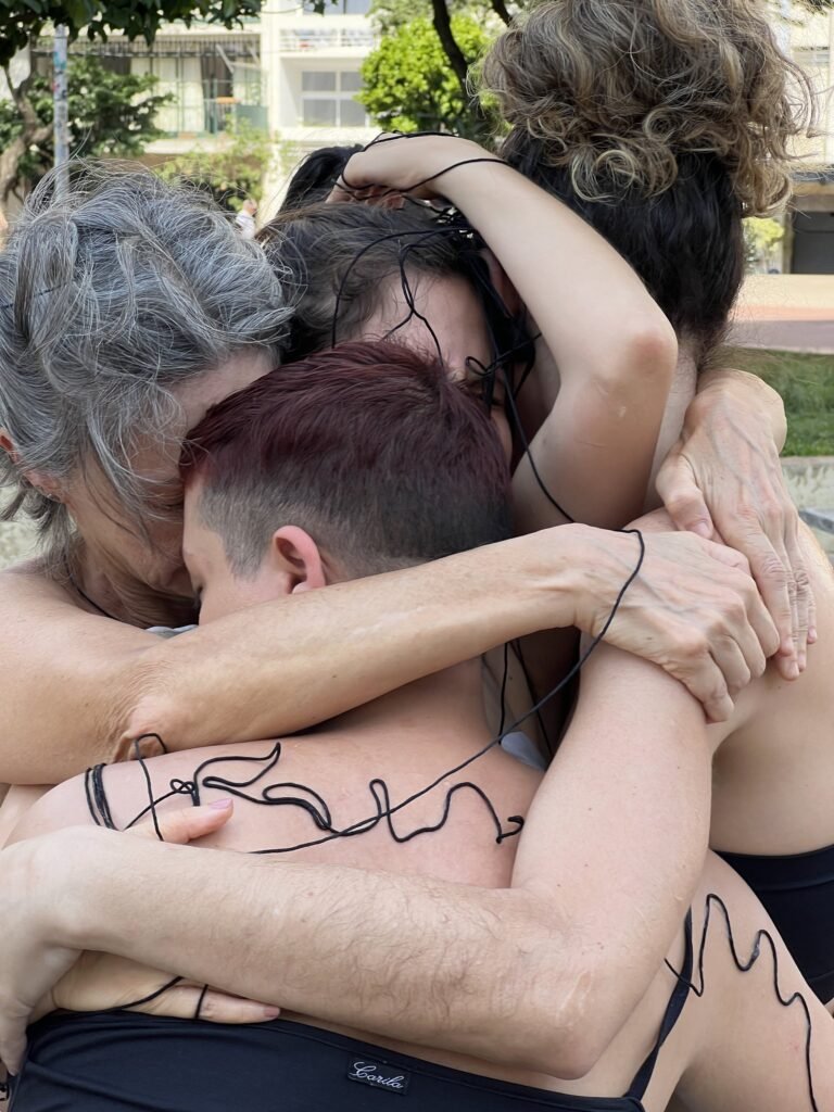 Performers se abraçam em grupo, com o fio preto desenhando palavras nas costas