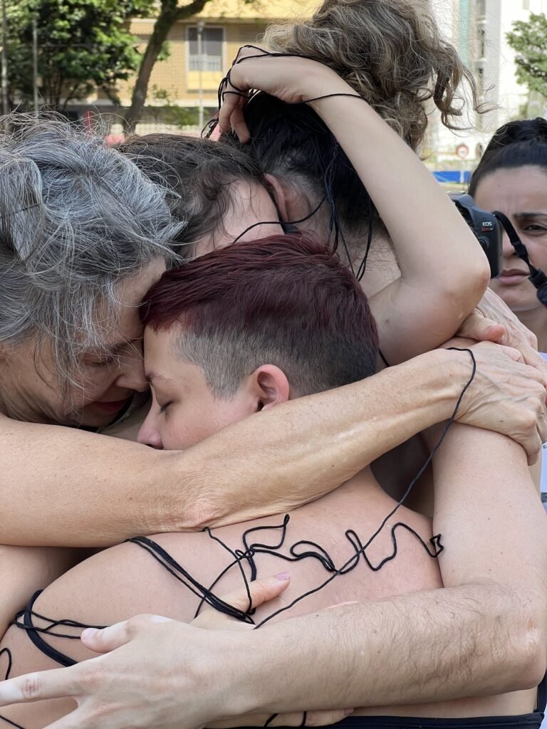 Abraço coletivo de sER.á.linha_do na Praça Roosevelt: cabeças encostadas e o fio preto escrevendo sobre as costas