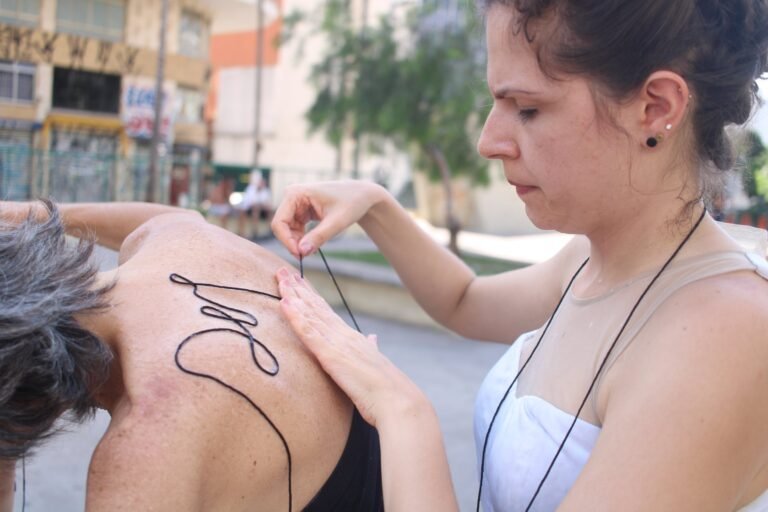 Uma performer passa o fio sobre as costas de outra, diante de um muro grafitado na Praça Roosevelt
