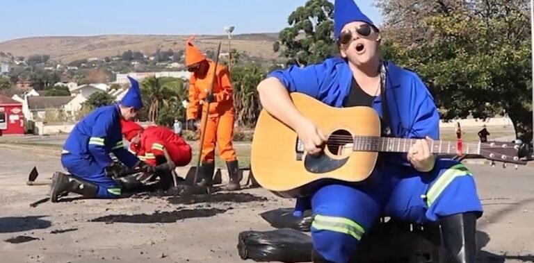 Fotograma de 12 Labours, de Gavin Krastin: artista vestido de gnomo toca violão ao lado de trabalhadores que consertam a rua