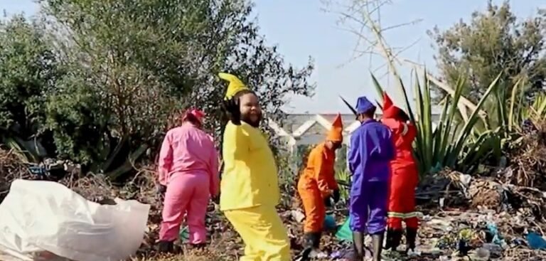 Fotograma de 12 Labours, de Gavin Krastin: artistas vestidos de gnomos, em roupas coloridas, num terreno com plantas