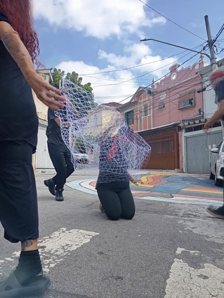 Pessoa coberta por uma rede ajoelhada na rua, com participantes em movimento em volta