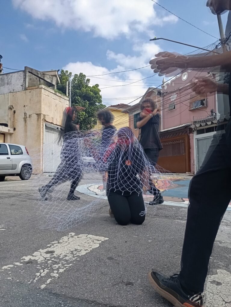 Participantes de preto se movem em volta de uma pessoa coberta por uma rede, numa rua do Bixiga