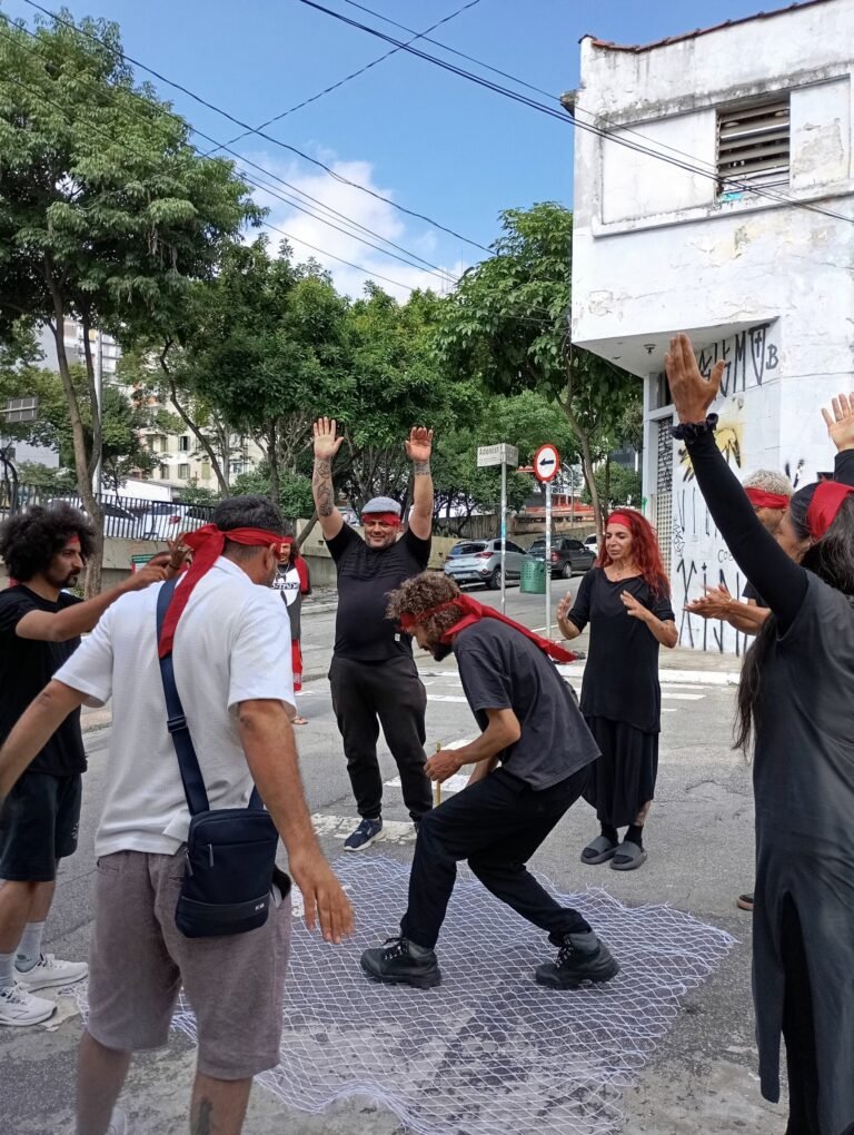Participantes de olhos vendados com tecidos vermelhos se movem numa rua do Bixiga