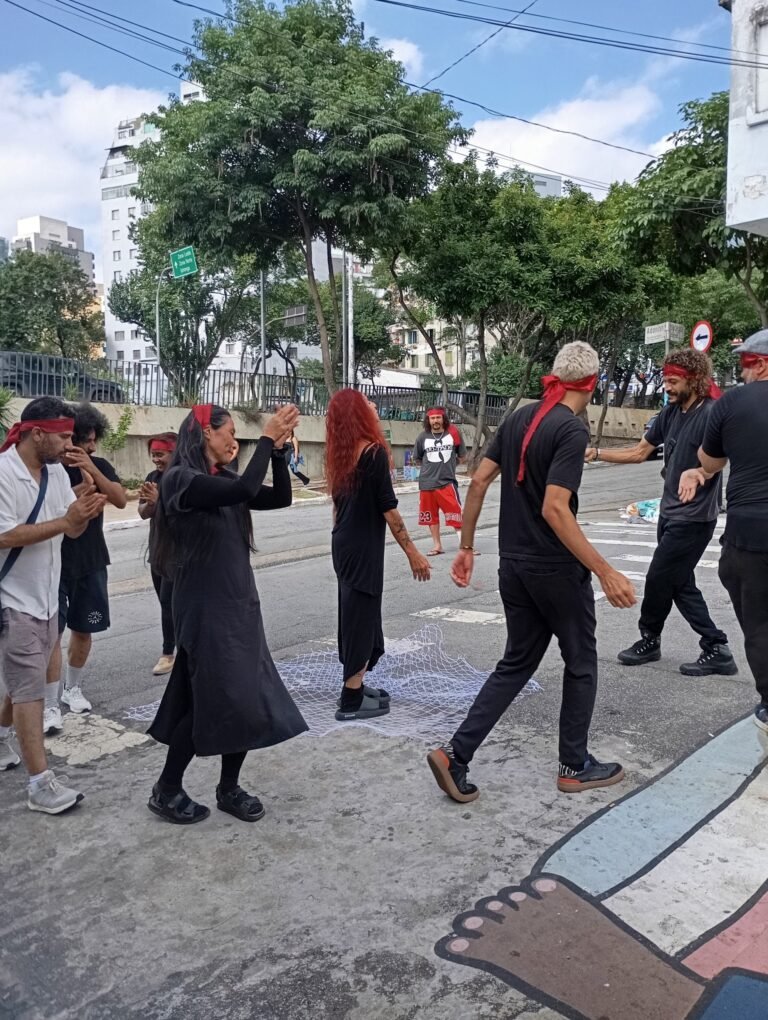 Participantes vestidos de preto com vendas vermelhas caminham em roda numa rua do Bixiga
