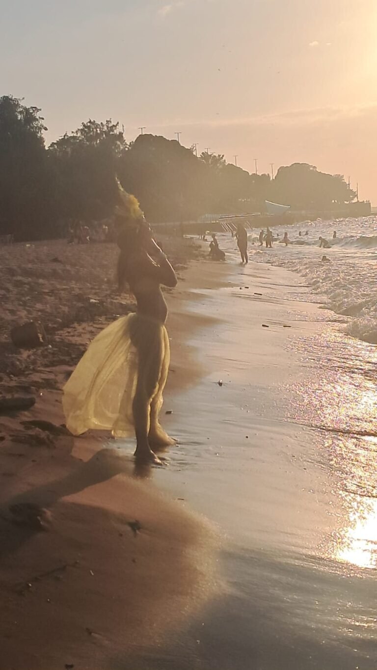 Icamiaba caminha na beira da água ao pôr do sol, de saia amarela