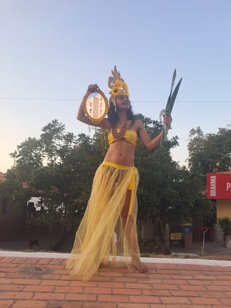 Icamiaba de saia de tule amarela ergue um espelho e folhas numa calçada de tijolos