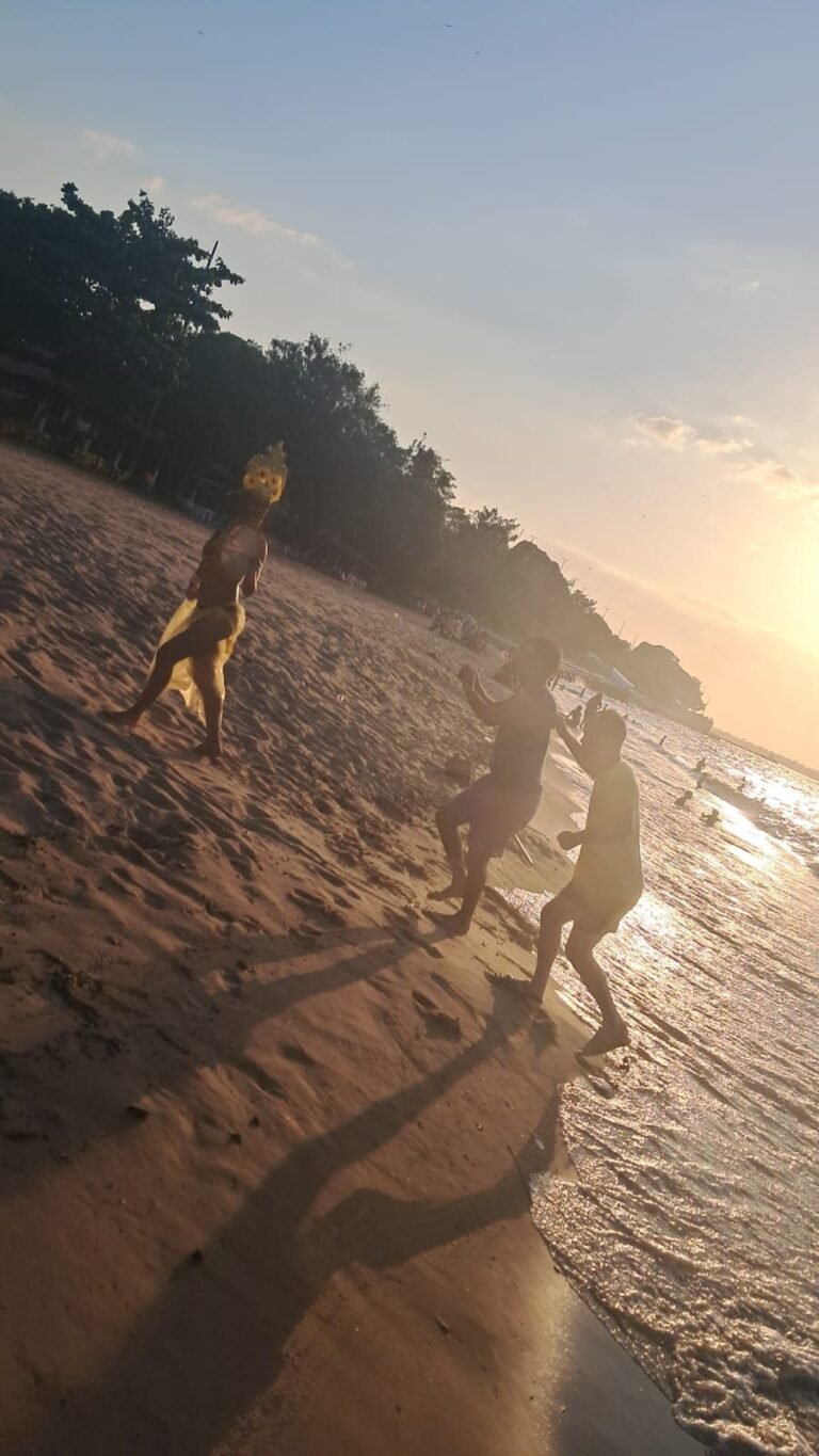 Icamiaba na areia, à beira da água, ao pôr do sol, com outras pessoas ao fundo