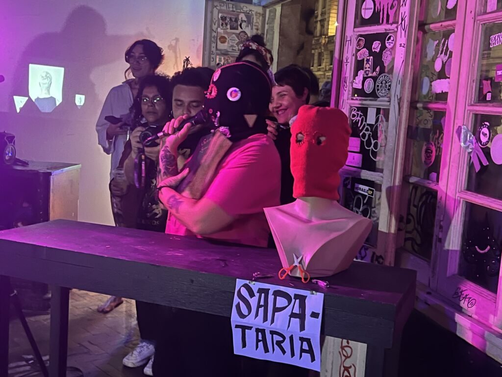 Integrantes da Boleia Coletiva atrás de uma bancada com a placa "Sapataria" e cabeças de manequim, no Estúdio Lâmina